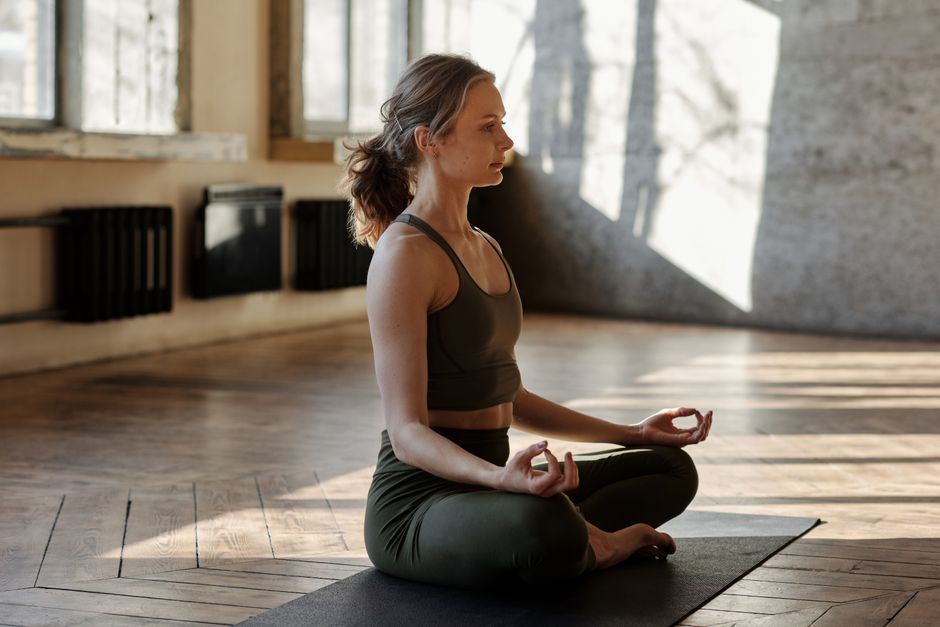 Woman sits in a meditative pose on a cushion in a bright, sun-drenched room, creating a feeling of peace and tranquility a...